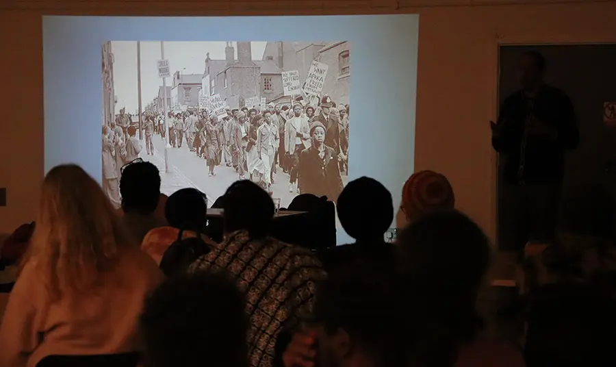 A group of people sit in a dark room watching a projection with images of a protest