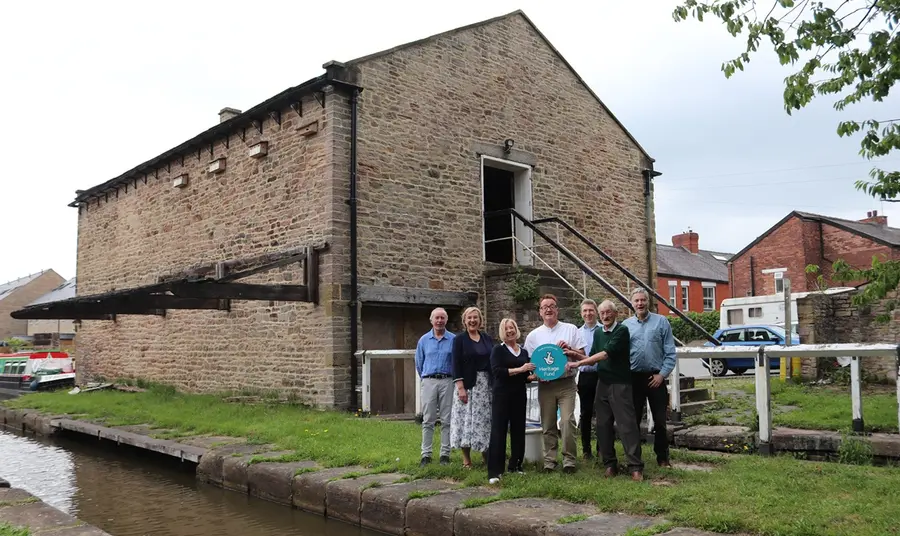 A group of smiling people, holding a blue Heritage Fund roundel, stand between the canal and the historic wharf building behind them.