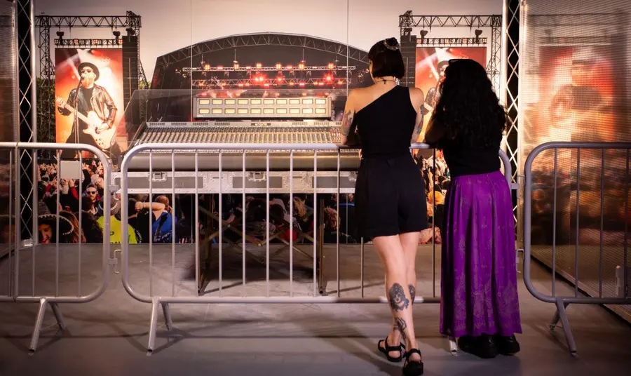 Two visitors stand at a festival-style barrier, looking at a large music mixing desk, decorated like a festival stage.