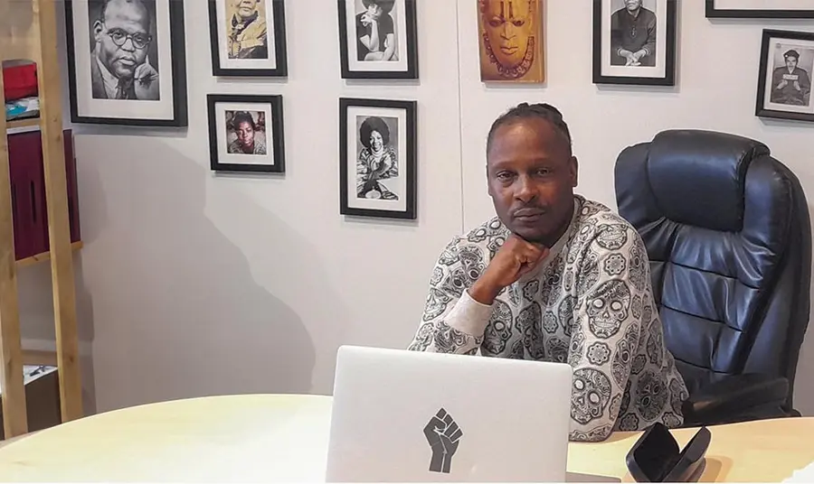 A black man wearing a smart patterned shirt sits behind a desk. On the wall behind him are framed photos of people of colour