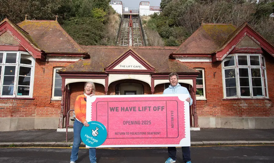 Two people stand holding a large ticket saying 'we have lift off' and bearing the Heritage Fund logo