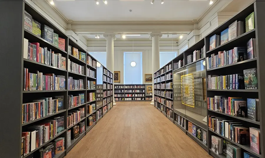 A broad aisle between two bookshelves in the refurbished library; a high ceilinged room, with large windows and classical columns.