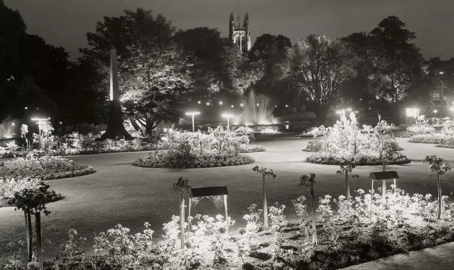 A black and white photograph of an elegant landscaped garden, illuminated. The tower of a church is illuminated in the background.