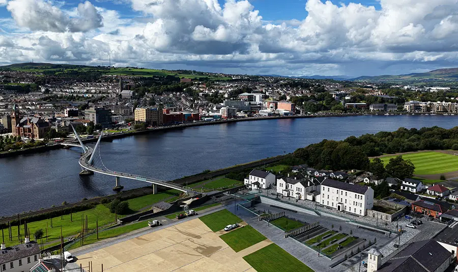 A public square with buildings next to it on the edge of a wide river, the city on the other side of the river is connected by a large white bridge