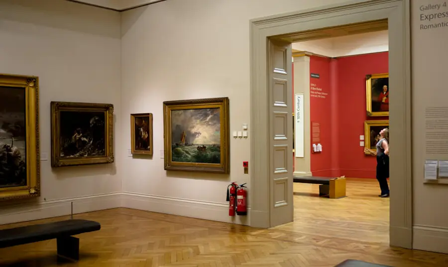 A gallery space with old paintings on the walls and a person in the background viewing the art