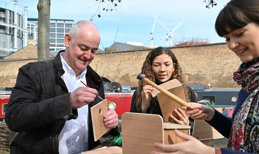 Three people sitting on a picnic table in an urban greenspace, building wooden birdboxes with hammers 