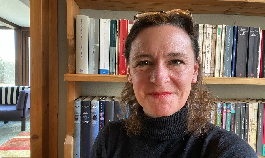 Caroline Crewe-Read A woman in a black polo neck jumper standing in front of a bookshel