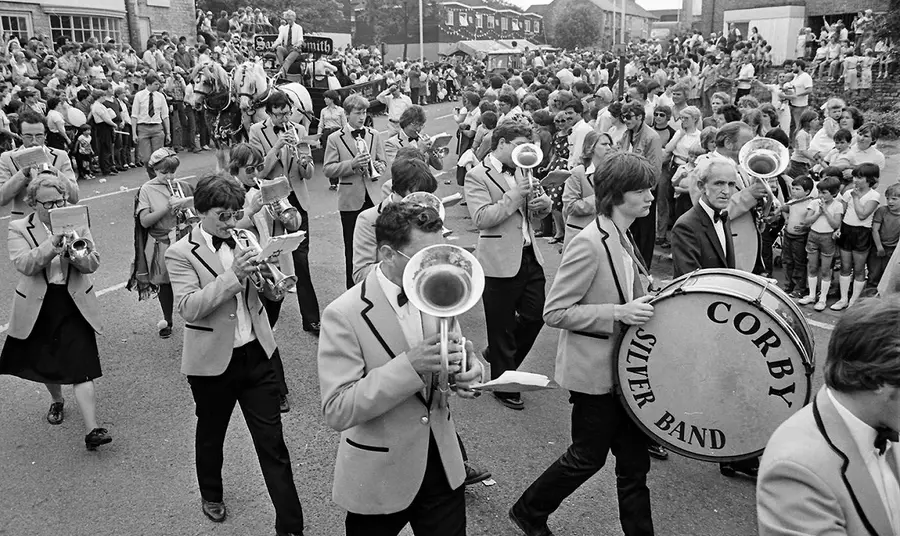 A black and white image of a brass band marching through the streets of a packed town centre.