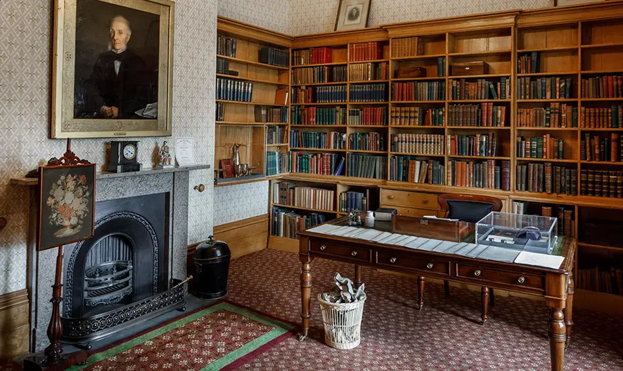 Inside a room with desk, book case and fire place in background