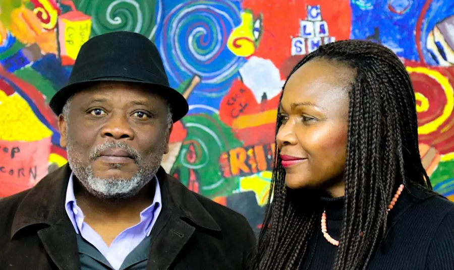 Oral History guidance Two people stand in front of a colourful wall, one looks at the other who is looking at the camera