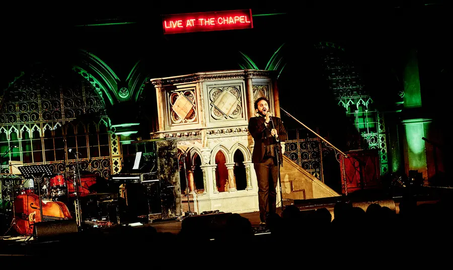 Nish Kumar performing at Union Chapel in front of pulpit and Live at the chapel LED sign