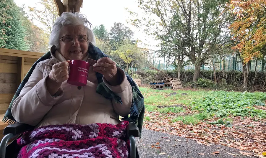 Elderly woman using wheelchair in garden, smiling with tea and biscuit
