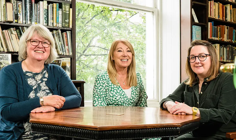 Three people sit at table in room surrounded by books on shelves. They smile at the camera.