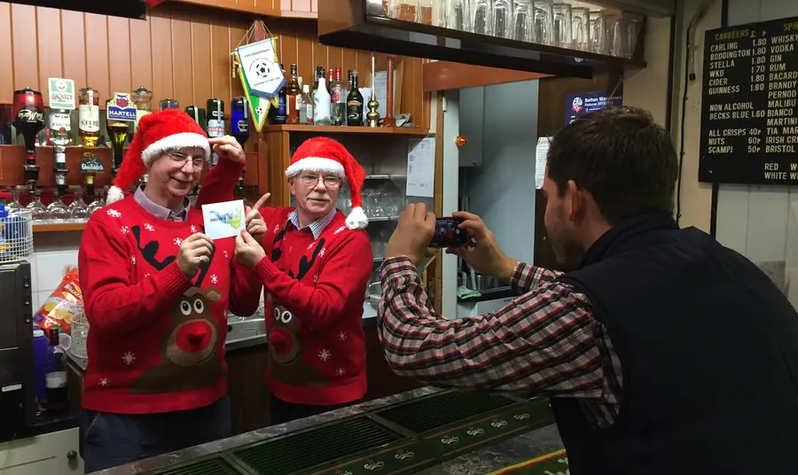 A person taking a photo of two people behind a bar wearing matching red Christmas hats and jumpers.