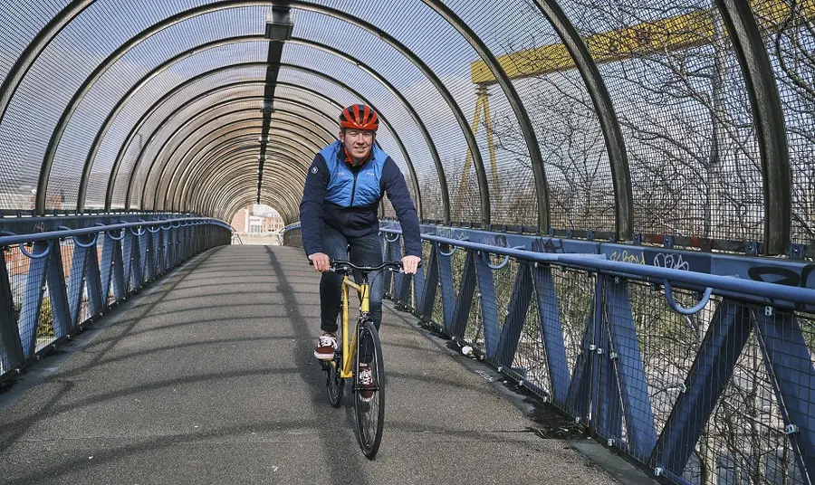 A man cycling on a bridge with the Harland and Wolff cranes visible in the background