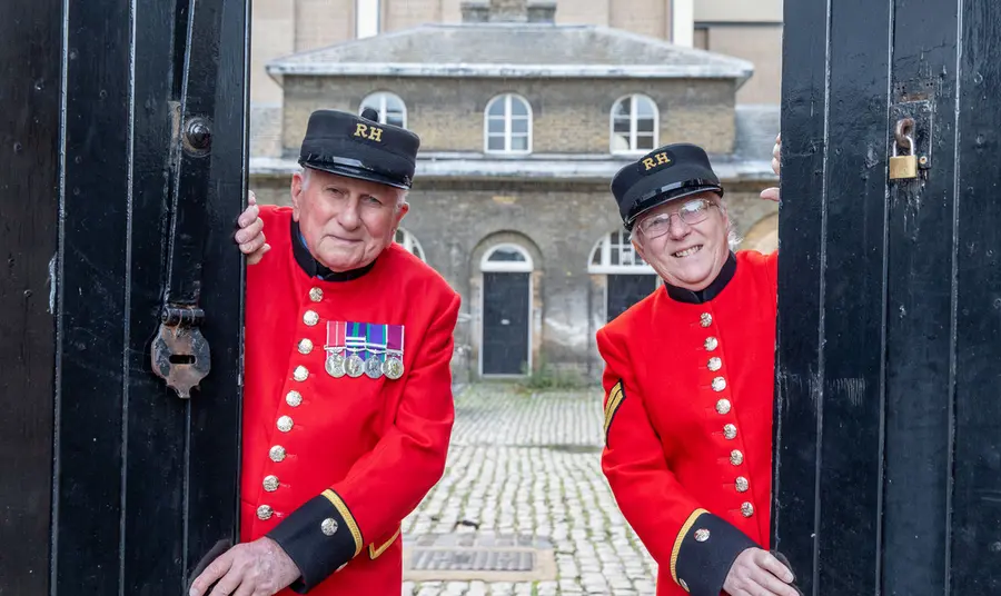 Two people wearing scarlet Chelsea Pensioners' uniform open stable doors at Royal Chelsea Hospital, London with historic building and courtyard behind