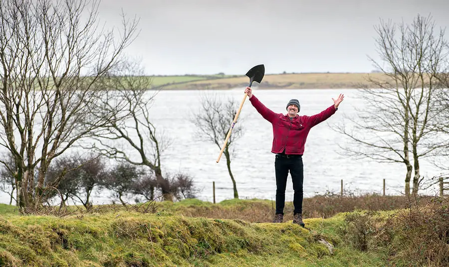 Person standing in nature looking happy whilst holding a spade in the air