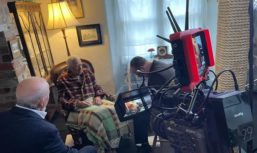 Behind the scenes at a film set: three actors act a scene in a living room while a camera films
