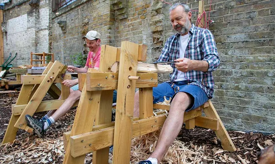 Two people sitting outside carving wood at woodworking benches