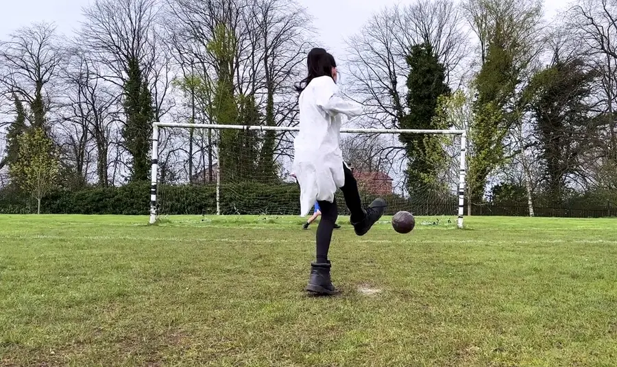 A young girl in a white dress and boots takes a penalty kick.