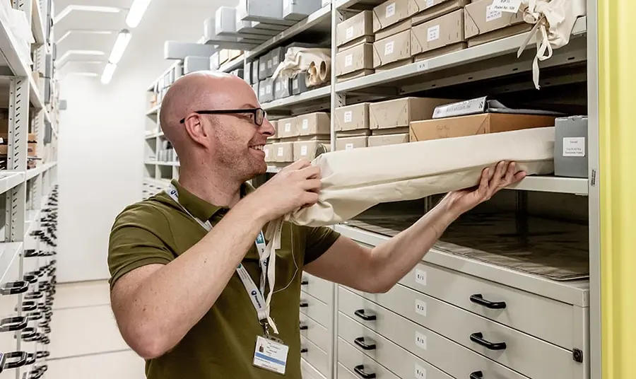 a man takes a cloth bag containing a rolled up document off a shelf in an archive