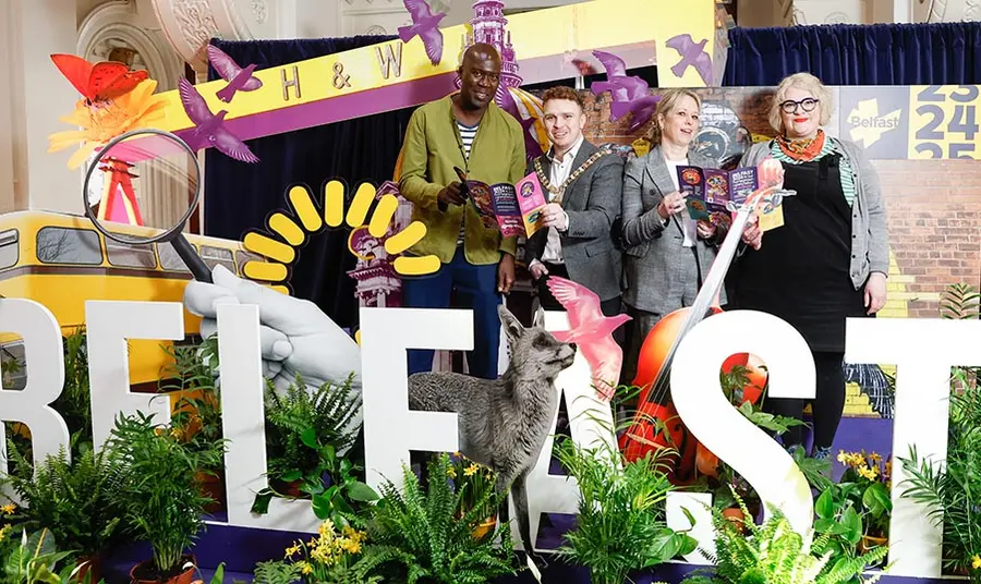 Four people pose with leaflets behind large letters spelling 'Belfast' surrounded by foliage and props