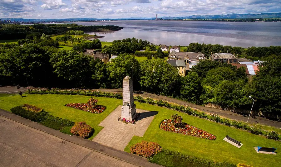 Bo'ness war memorial is at the centre of the frame and behind it there are trees and houses. In the distance is the Firth of Forth.