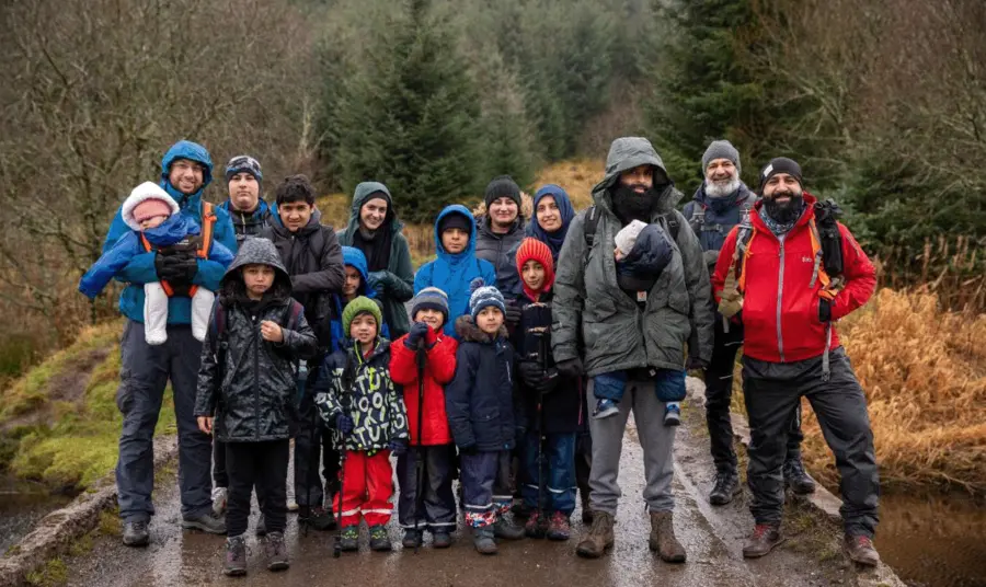 A large group of people of all ages in walking gear on a rainy day. They are standing on a path through a wooded glen.