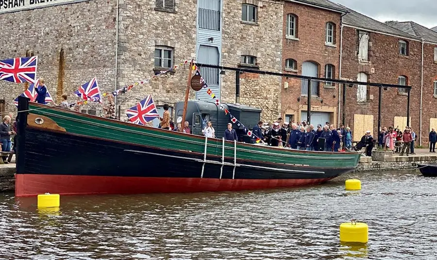 A crowd of people, some in maritime uniforms, gathered around a ship's hull docked next to harbour buildings. The ship is flying Union Jack flags and nautical bunting.