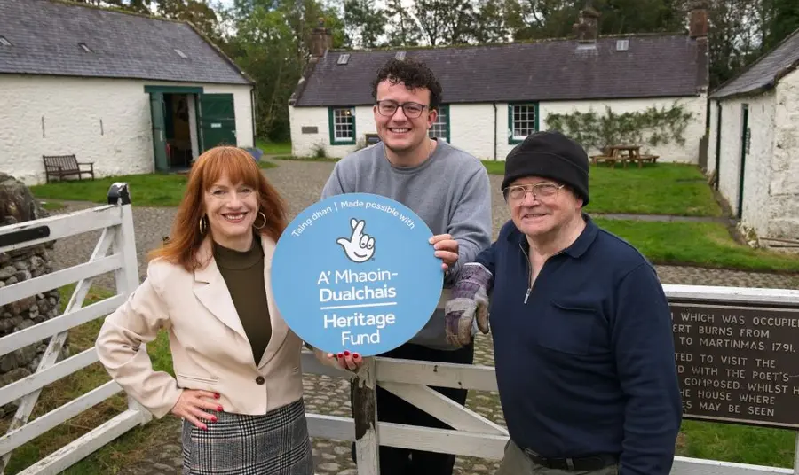 Three people standing at a gate. Behind them are white farm buildings. They are holding a blues circular The NAtional Lottery Heritage FUnd sign