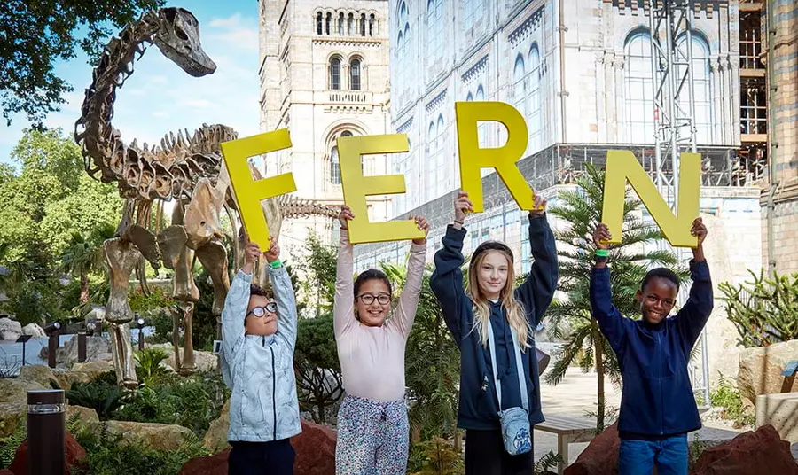 four children hold up large letters spelling FERN, the name of the diplodocus skeleton on display behind them
