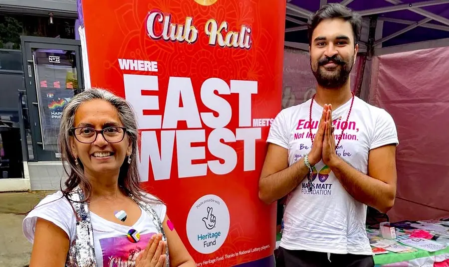 Two south asian people stand next to a banner with text: Club Kali Where East Meets West made possible with Heritage Fund