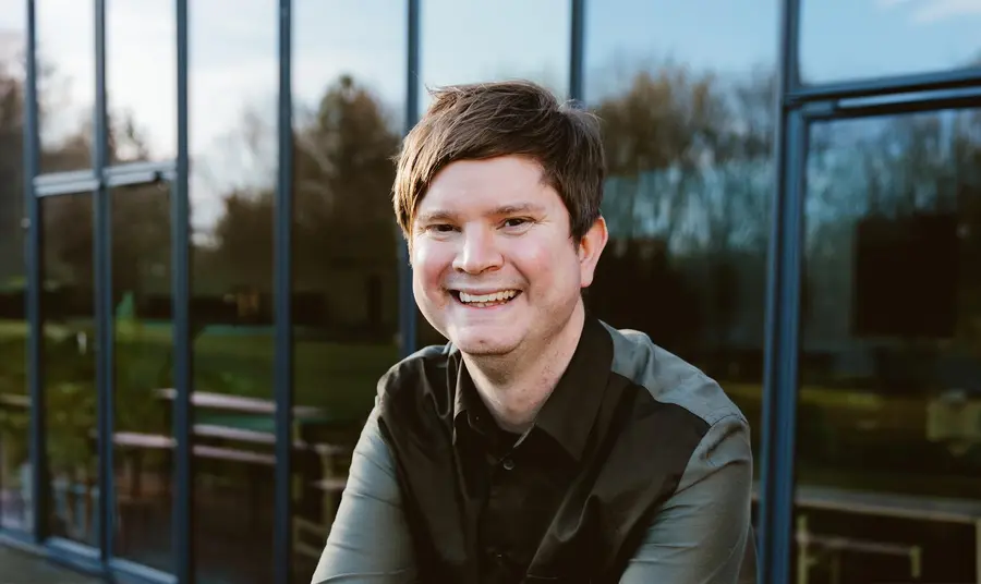 a photo of David Sheppeard sitting outside against a glass wall. David has white skin, short brown hair and wears a dark shirt