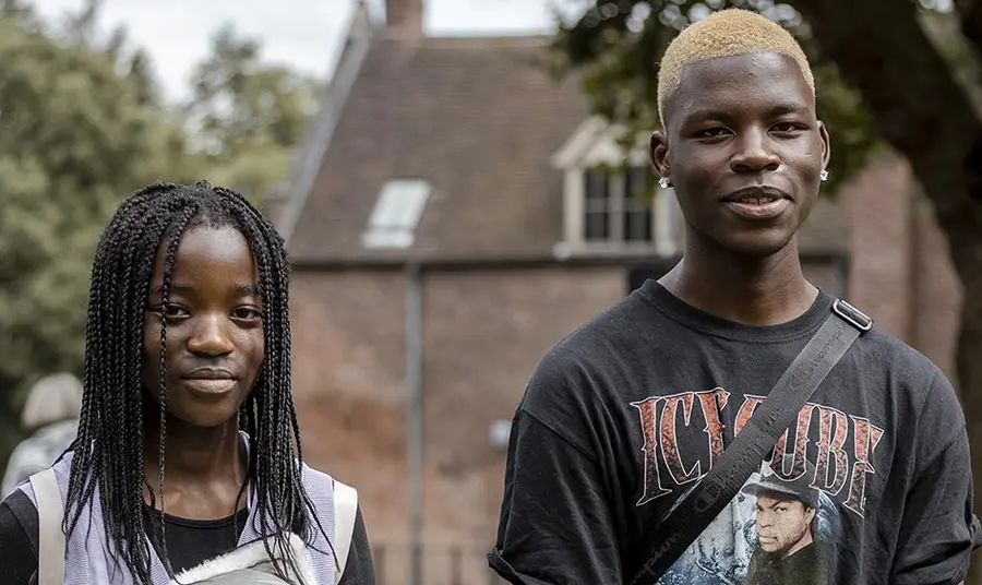 Two young black people wearing casual clothes stand outside with a historic brick building in the background