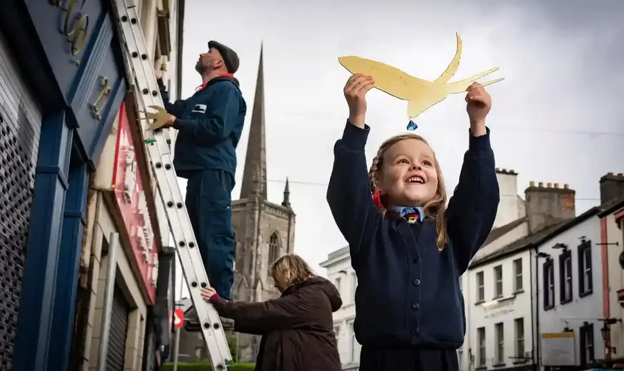 A young girl holds a cut out of a golden bird above her head. In the background are two adults working on a ladder.