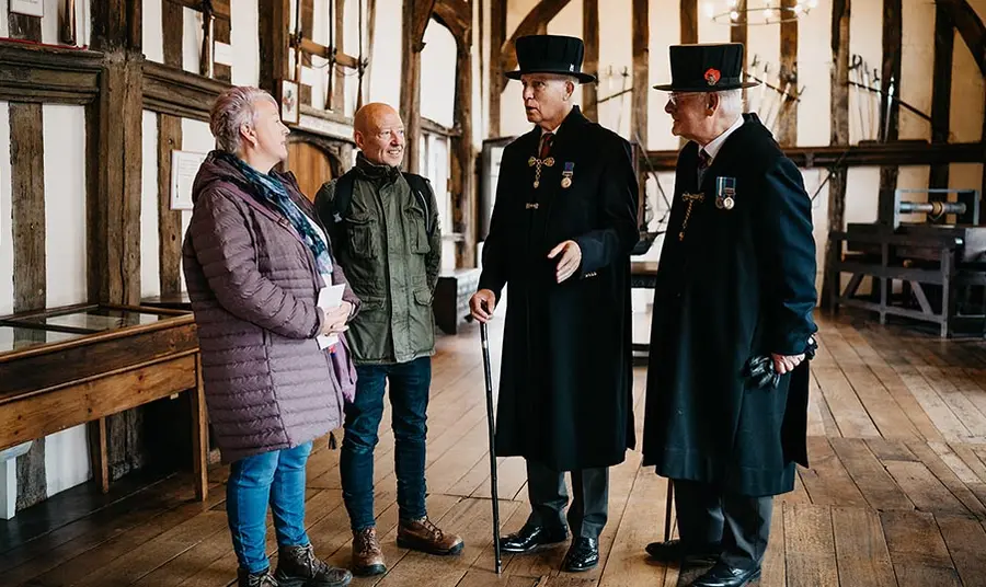 two visitors are given a tour of a timber framed hall by two older gentlemen wearing long black coats at traditional hats