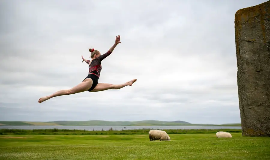 A gymnast leaps through the air in a field on Orkney, with sheep and standing stones nearby.