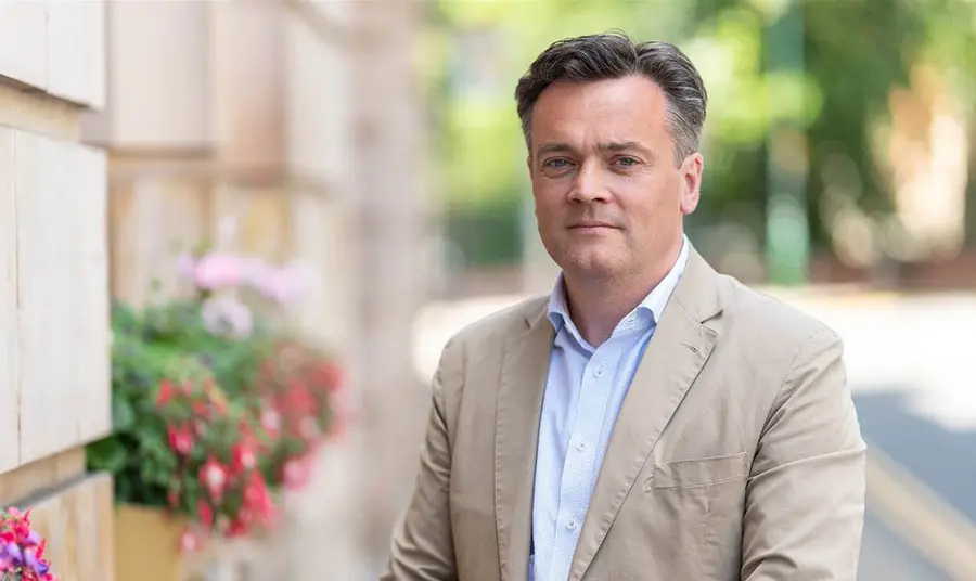 James Twining wearing shirt and jacket, standing next to a stone building with flower boxes.