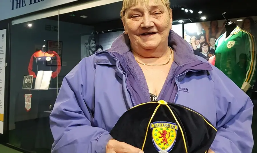 Janie Houghton, History Maker A woman in a purple jacket holding a Scotland international football cap with the name Janie Houghton embroidered on the peak