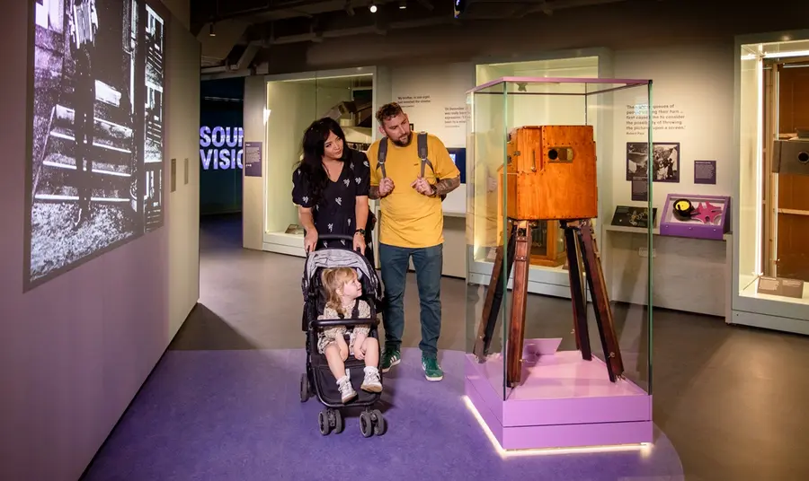 A man and a woman push a child in a buggy as they take a closer look at a pioneering film camera from the early days of the technology. A black and white film of the era is projected on the wall to their left.