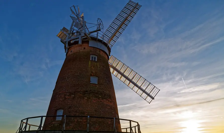 John Webb's windmill at Thaxted, Essex. Looking up at a red brick windmill with large white sails, as the sun sets to the right.