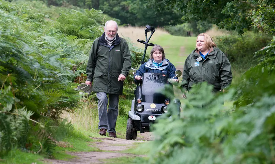 A man and woman walking alongside a woman in a mobility scooter on a path surrounded by plants at RSPB Minsmere