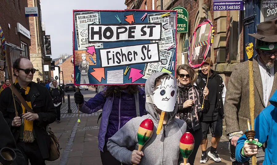 a parade in a town high street, one person holds up a sign with 'hope street' on it and a young person in a homemade mask shakes maracas