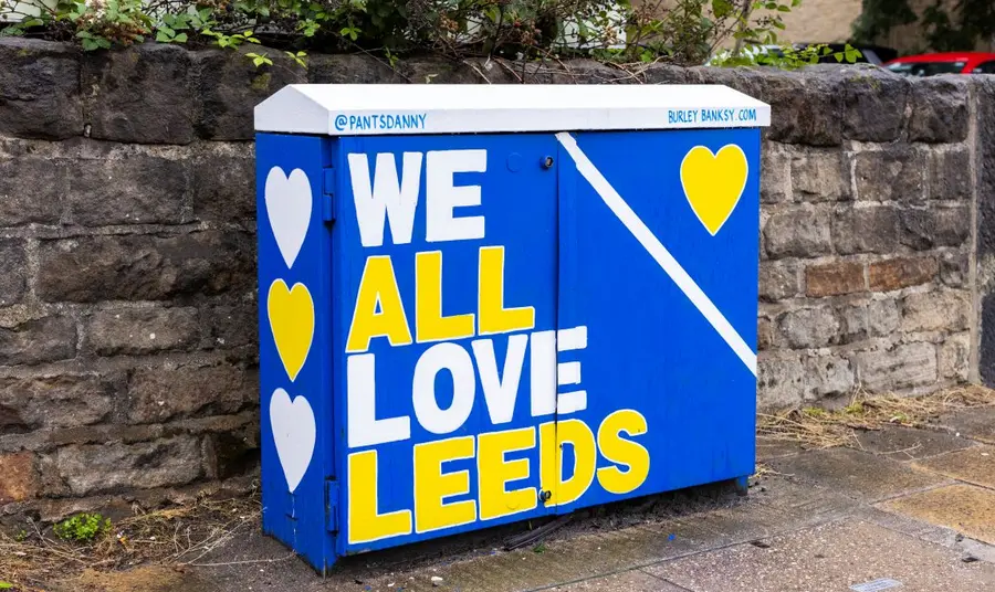 leedm.e.2023.0034.0066_burley_banksy.jpg A telephone cable cabinet decorated in blue and yellow, with the slogan 'We All Love Leeds'.