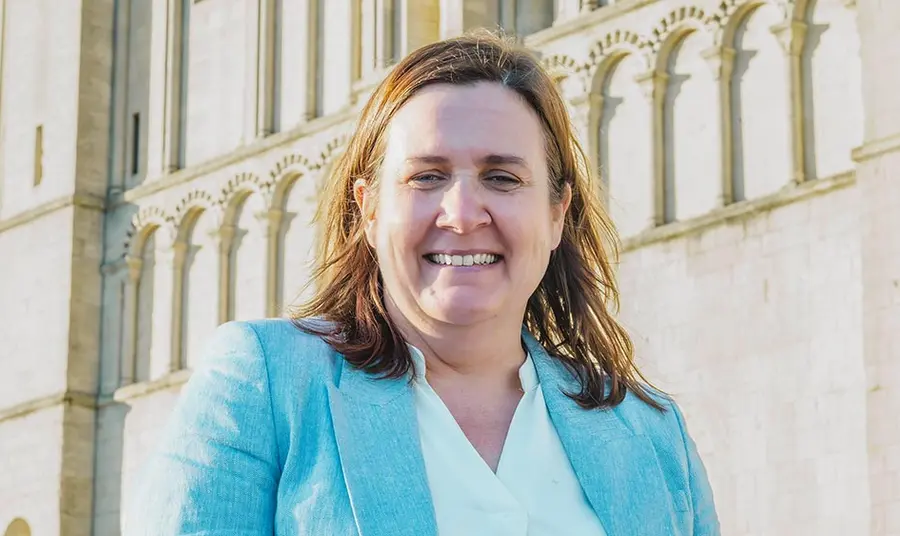 A head and shoulders photo of Liz Bates, a woman with fair skin and brown hair wearing a suit, standing in front of Norwich Castle