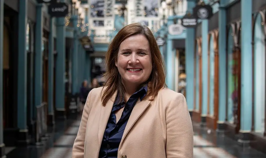 A photo of Liz Bates, a woman with fair skin and brown hair wearing a blue silk shirt and cream coat, standing in a historic shopping arcade