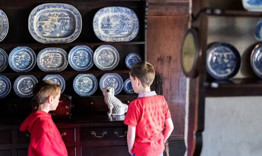 Children visit St. Fagans Museum, Wales. Two children, facing away, looking at a wall of traditional porcelain plates.