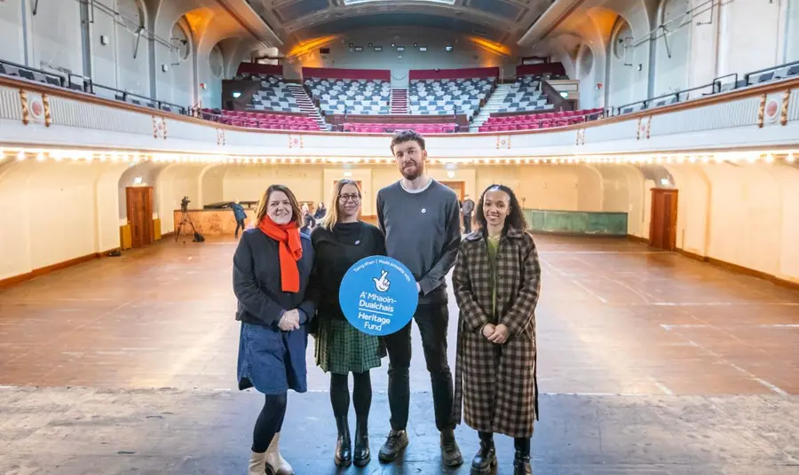 Funds have been awarded to Leith Theatre Trust. A group of four colleagues, holding up a blue Heritage Fund roundel, stand in the middle of the stage of the Leith Theatre, looking out to the empty stalls and grand circle above.