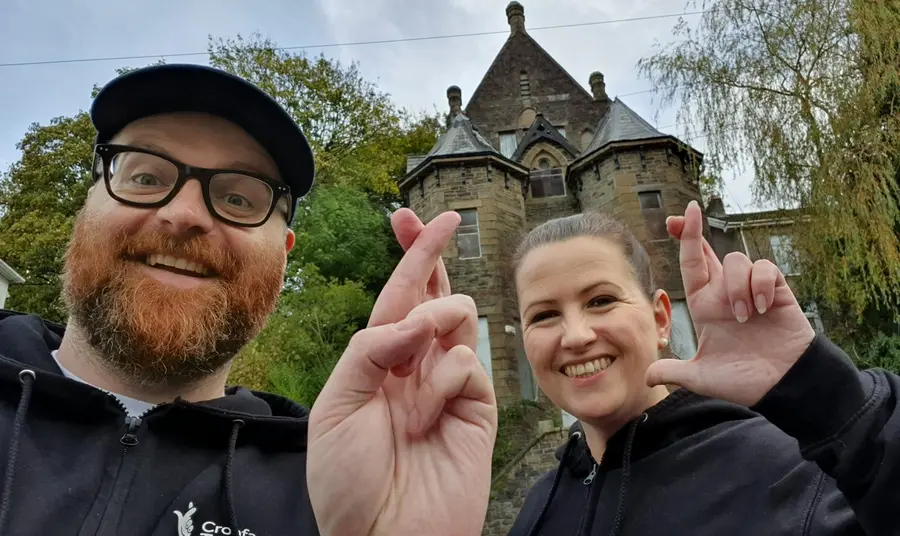 A man and a woman are stood in front of a heritage site doing the crossed fingers selfie