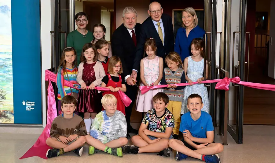 A group of children and adults sit and stand as they pose for a photo while cutting a ribbon.